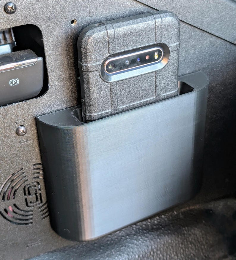 Puede incluir: Una funda de tel&eacute;fono negra con un tel&eacute;fono dentro se encuentra en un soporte de tel&eacute;fono de pl&aacute;stico negro montado en un coche.