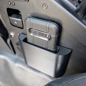Puede incluir: Una funda de tel&eacute;fono negra con un soporte de tel&eacute;fono integrado, dise&ntilde;ada para caber en el interior de un veh&iacute;culo. La funda del tel&eacute;fono est&aacute; montada en un soporte de pl&aacute;stico negro, que est&aacute; unido al lado del asiento de un veh&iacute;culo.
