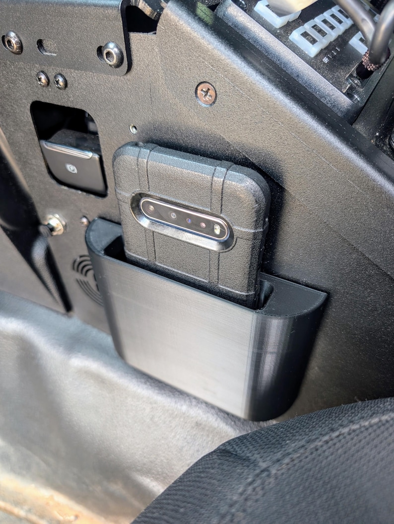 Puede incluir: Una funda de tel&eacute;fono negra con un soporte de tel&eacute;fono integrado, dise&ntilde;ada para caber en el interior de un veh&iacute;culo. La funda del tel&eacute;fono est&aacute; montada en un soporte de pl&aacute;stico negro, que est&aacute; unido al lado del asiento de un veh&iacute;culo.