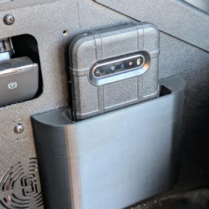 Puede incluir: Un soporte de tel&eacute;fono negro est&aacute; montado en la consola central de un coche. El soporte est&aacute; hecho de pl&aacute;stico y tiene un borde redondeado. Un tel&eacute;fono negro con una lente de c&aacute;mara est&aacute; en el soporte.