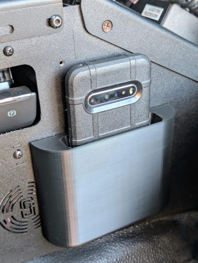 Puede incluir: Un soporte de tel&eacute;fono negro est&aacute; montado en la consola central de un coche. El soporte est&aacute; hecho de pl&aacute;stico y tiene un borde redondeado. Un tel&eacute;fono negro con una lente de c&aacute;mara est&aacute; en el soporte.