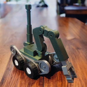 May include: A green and silver toy robot with a robotic arm and wheels. The robot is on a wooden surface.