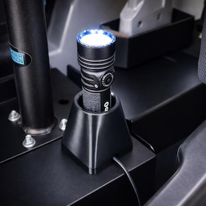 May include: A black flashlight with a bright white light and blue accents is in a black charging base. The flashlight has a textured grip and a control dial. A charging cable is connected to the base.
