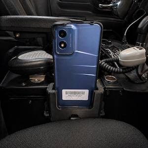 May include: A blue smartphone in a black case is secured in a black holder. The phone has dual cameras and a barcode sticker. The holder is mounted in a vehicle's interior, near a microphone and other controls.