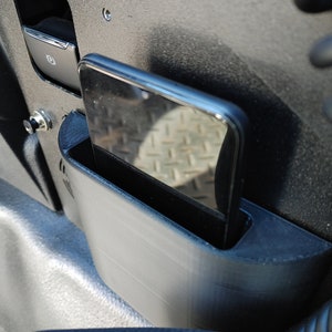 Puede incluir: Un tel&eacute;fono negro se coloca en un soporte de tel&eacute;fono gris que est&aacute; unido al lado de un asiento de coche negro.