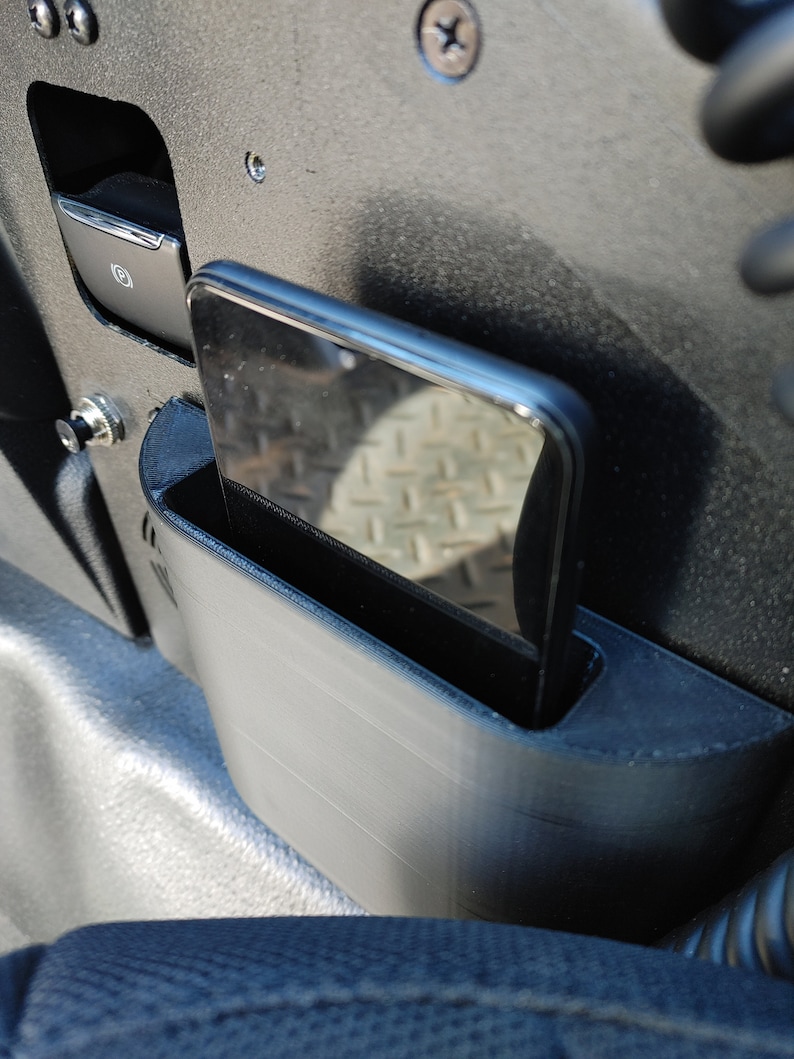 Puede incluir: Un tel&eacute;fono negro se coloca en un soporte de tel&eacute;fono gris que est&aacute; unido al lado de un asiento de coche negro.