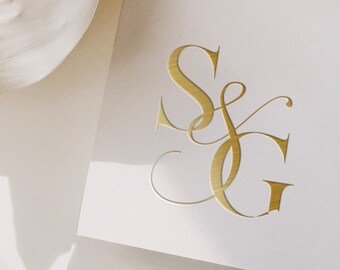 May include: A white card with gold-colored initials "SG" in an elegant script. The card is next to white, feathery floral elements. The image has soft lighting and a minimalist aesthetic, suitable for wedding stationery or event announcements.