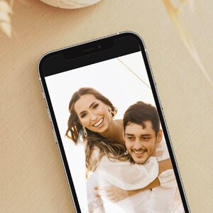 May include: A close-up photo of a couple smiling at the camera. The woman is wearing a white dress and has her arms around the man's neck. The man is wearing a white shirt and is looking at the camera. The photo is displayed on a smartphone.