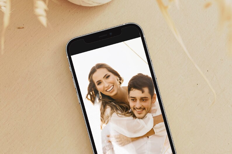 May include: A close-up photo of a couple smiling at the camera. The woman is wearing a white dress and has her arms around the man's neck. The man is wearing a white shirt and is looking at the camera. The photo is displayed on a smartphone.