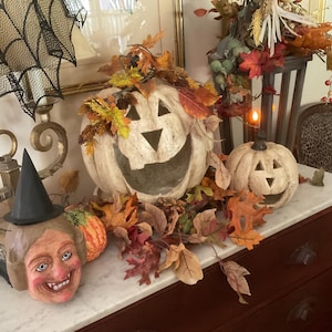 May include: A collection of Halloween decor featuring two off-white carved pumpkins with triangular eyes and mouths, a witch head figurine wearing a black pointed hat, and a decorative arrangement of artificial autumn leaves and berries.