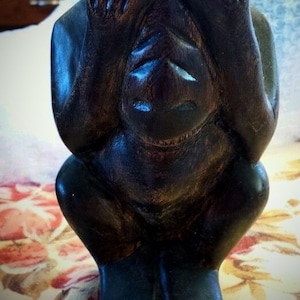 May include: A dark wood carving of a monkey covering its eyes with its hands. The monkey is sitting on a floral patterned surface.
