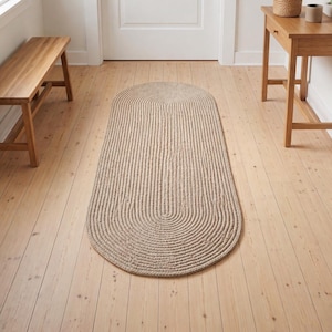 Pode incluir: Um tapete bege, de formato oval, com um design texturizado e canelado. O tapete está colocado sobre um chão de madeira clara, e o design apresenta uma série de linhas verticais que se curvam na parte inferior. É um artigo de decoração para casa.