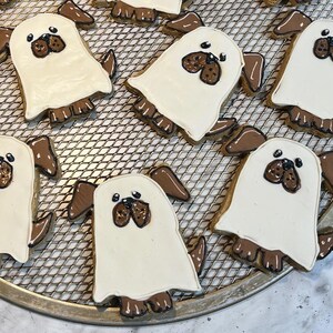 May include: Several dog-shaped cookies with white icing and brown details are arranged on a metal cooling rack. The cookies have a cartoon-like appearance, with brown ears, noses, and paws. The cookies are baked goods.