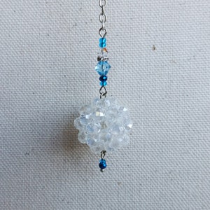 May include: A clear, spherical beaded ornament with a cluster of faceted beads. The ornament is suspended by a silver chain, with blue and clear beads accenting the chain. A small blue bead dangles at the bottom.