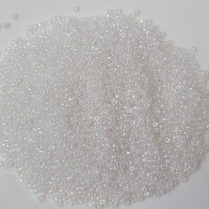 May include: A large pile of small, clear iridescent seed beads. The beads are scattered on a white surface.