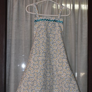 May include: A light blue and white daisy print apron with a turquoise trim along the top edge. The apron is hanging on a white hanger against a sheer curtain background.