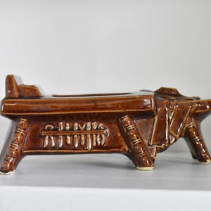 May include: A brown ceramic planter shaped like a small bench with decorative details. The planter has a rectangular base and two legs on each side.