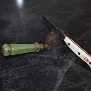 Puede incluir: Un afilador de cuchillos con un mango de madera verde y un afilador de cuchillas de acero inoxidable. Un cuchillo con mango blanco y remaches negros descansa en el afilador. El cuchillo y el afilador est&aacute;n sobre una superficie oscura.