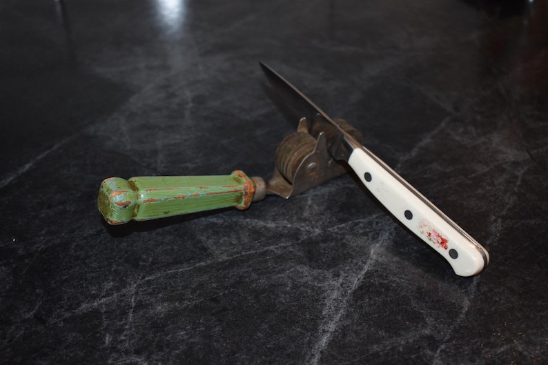 Puede incluir: Un afilador de cuchillos con un mango de madera verde y un afilador de cuchillas de acero inoxidable. Un cuchillo con mango blanco y remaches negros descansa en el afilador. El cuchillo y el afilador est&aacute;n sobre una superficie oscura.