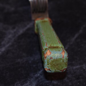 Puede incluir: Primer plano de un tenedor con un mango de madera verde desgastado. El mango muestra signos de desgaste con madera expuesta y pintura descolorida. Se ven las p&uacute;as met&aacute;licas del tenedor.