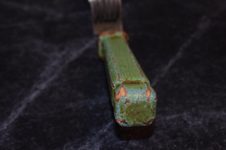 Puede incluir: Primer plano de un tenedor con un mango de madera verde desgastado. El mango muestra signos de desgaste con madera expuesta y pintura descolorida. Se ven las p&uacute;as met&aacute;licas del tenedor.