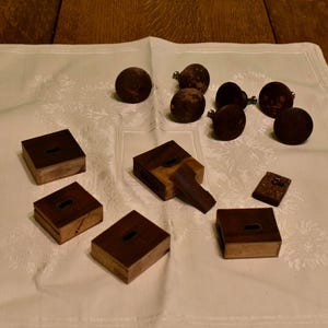 Puede incluir: Un surtido de pequeñas cajas de madera marrón oscuro y objetos redondos con tallos, dispuestos sobre un paño blanco con un estampado floral. Las cajas son rectangulares con aberturas en forma de ojo de cerradura. Los objetos redondos son marrón oscuro.
