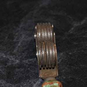 Puede incluir: Primer plano de una herramienta met&aacute;lica vintage con m&uacute;ltiples ruedas ranuradas. La herramienta tiene un aspecto oxidado y envejecido con un mango de madera pintado de verde y naranja. Las ruedas se sujetan con peque&ntilde;os remaches met&aacute;licos.