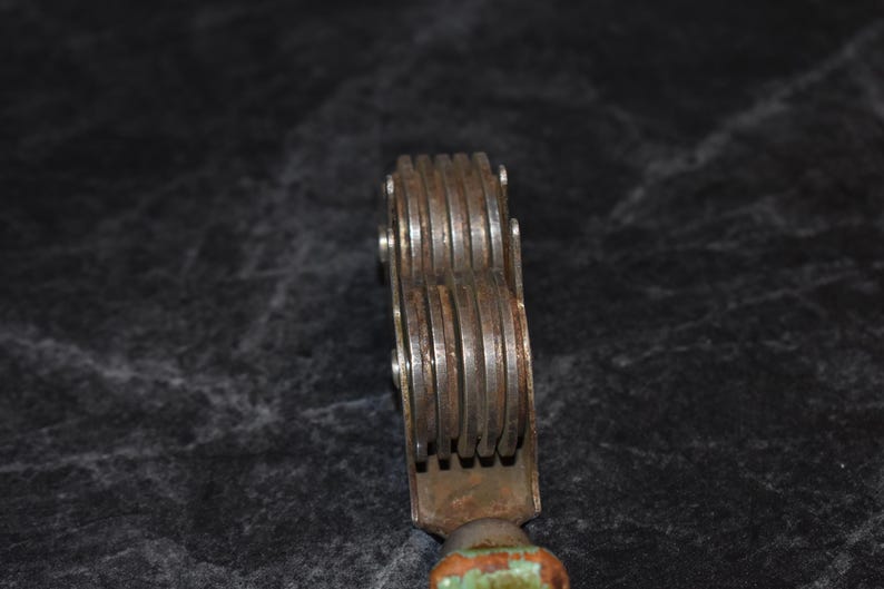 Puede incluir: Primer plano de una herramienta met&aacute;lica vintage con m&uacute;ltiples ruedas ranuradas. La herramienta tiene un aspecto oxidado y envejecido con un mango de madera pintado de verde y naranja. Las ruedas se sujetan con peque&ntilde;os remaches met&aacute;licos.
