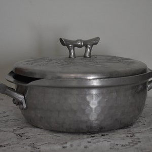 May include: A silver hammered aluminum casserole dish with a lid and bamboo-style handles. The dish has a floral design on the lid.