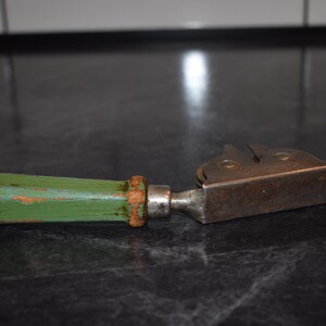 Puede incluir: Un abrelatas antiguo con un mango de madera pintado de verde y una cuchilla de metal. El mango muestra signos de desgaste, con la pintura verde desconchada en algunos lugares. La cuchilla de metal tiene un aspecto vintage y r&uacute;stico.