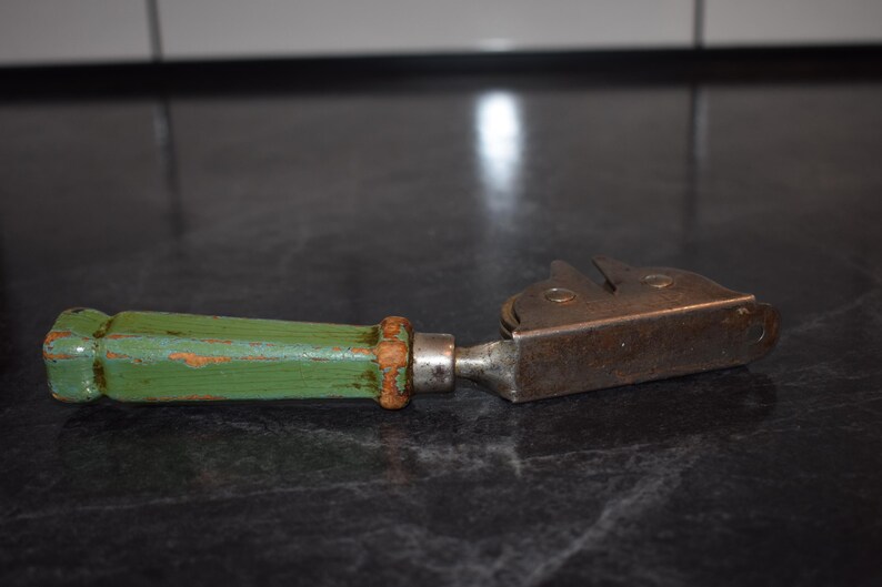Puede incluir: Un abrelatas antiguo con un mango de madera pintado de verde y una cuchilla de metal. El mango muestra signos de desgaste, con la pintura verde desconchada en algunos lugares. La cuchilla de metal tiene un aspecto vintage y r&uacute;stico.