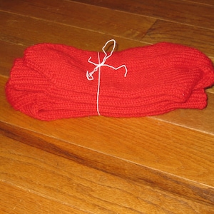 May include: A stack of bright red knitted items, possibly socks or a scarf, tied together with white string. The items are made of a thick, textured yarn and are neatly folded. The background is a wooden floor.