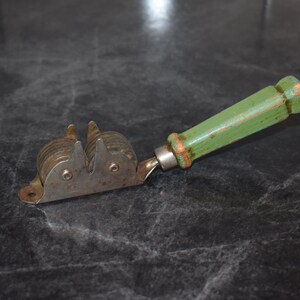 Puede incluir: Un afilador de cuchillos vintage con un mango de madera verde y un mecanismo de afilado de metal. El mango tiene un aspecto desgastado y los componentes met&aacute;licos muestran signos de uso. El afilador est&aacute; dise&ntilde;ado para uso manual.