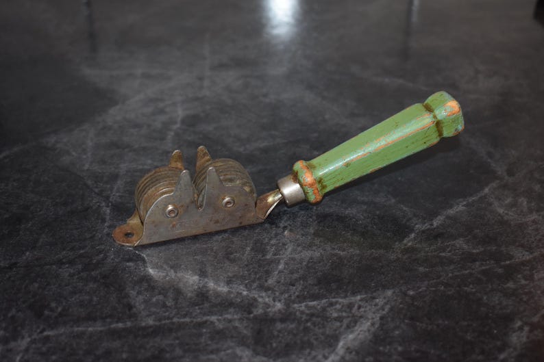 Puede incluir: Un afilador de cuchillos vintage con un mango de madera verde y un mecanismo de afilado de metal. El mango tiene un aspecto desgastado y los componentes met&aacute;licos muestran signos de uso. El afilador est&aacute; dise&ntilde;ado para uso manual.