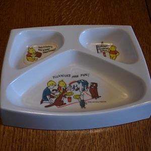 Può includere: Un piatto diviso in tre sezioni di colore blu con Winnie the Pooh. La sezione centrale ha un'immagine di Winnie the Pooh e i suoi amici che fanno un picnic con il testo "Picknicks are fun!" La sezione in alto a sinistra ha un'immagine di Winnie the Pooh con il testo "Does it funny how a bear likes honey?" La sezione in alto a destra ha un'immagine di Winnie the Pooh con il testo "But he's interrupted by Piglet."