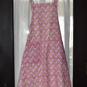May include: A pink and white chevron patterned apron with yellow accents, hanging on a wooden hanger. The apron has a square neckline, shoulder straps, and a tie at the waist. The fabric is a light cotton.