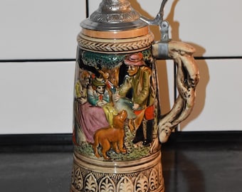 Puede incluir: Una jarra de cerveza de cerámica decorativa con una escena en relieve detallada de personas y un perro. La jarra tiene una tapa y un asa de metal. Los colores incluyen beige, marrón, verde y toques de rojo y azul.