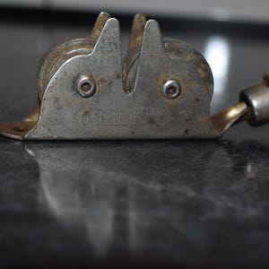 Puede incluir: Afilador de cuchillos de metal vintage con mango verde. El afilador tiene dos muelas y una gu&iacute;a para la cuchilla. El metal presenta un aspecto desgastado y envejecido. El mango es verde con signos de uso. Una herramienta pr&aacute;ctica para la cocina.