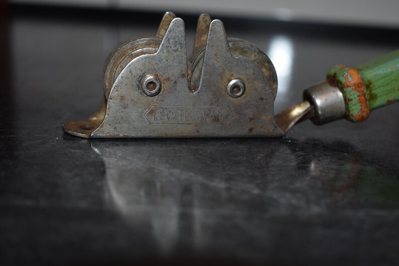 Puede incluir: Afilador de cuchillos de metal vintage con mango verde. El afilador tiene dos muelas y una gu&iacute;a para la cuchilla. El metal presenta un aspecto desgastado y envejecido. El mango es verde con signos de uso. Una herramienta pr&aacute;ctica para la cocina.