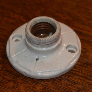 Puede incluir: Un portalámparas de cerámica blanca vintage con base circular y abertura central para una bombilla. El portalámparas tiene dos orificios de montaje y una superficie texturizada. El interior es de color marrón oscuro.