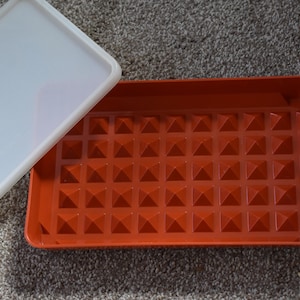 Puede incluir: Una bandeja de plástico roja con una superficie texturizada de pequeñas pirámides. La bandeja tiene una tapa de plástico blanco.