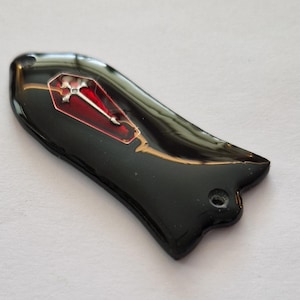 Puede incluir: Un objeto decorativo negro brillante con forma de ataúd. El ataúd presenta un interior rojo y una cruz plateada. La pieza tiene una forma curva y un agujero, posiblemente para un colgante o amuleto.