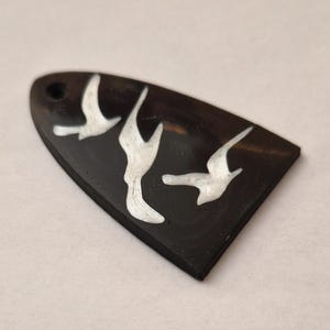 May include: A black, shield-shaped pendant with three white bird silhouettes. The pendant has a small hole at the top for hanging. The birds are in flight, adding a sense of movement to the design.