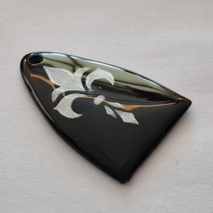 May include: A black, teardrop-shaped pendant with a white fleur-de-lis design. The pendant has a glossy finish and a small hole at the top for a chain or cord. The design is a classic symbol.