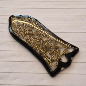 Può includere: Un pezzo unico in resina fatto a mano con un bordo nero. La resina trasparente racchiude numerosi piccoli chiodi color ottone, creando un effetto visivo sorprendente. La forma è allungata con un bordo decorativo.