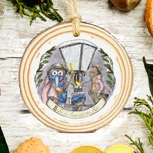 May include: A round wooden ornament with a hand-drawn illustration of two Muppet characters near a street lamp. The ornament has a natural wood grain edge and a jute twine hanger. The text on the banner reads "Light the lamp not the Rat."