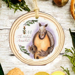 Puede incluir: Adorno navideño redondo de madera con una ilustración de un armadillo. El armadillo tiene un cuerpo marrón y un caparazón estampado, sobre un fondo morado claro. El texto "The Holiday Armadillo" está en el adorno.