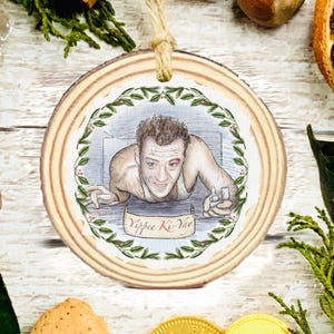 May include: A circular wooden Christmas decoration with a watercolour illustration of a man with a black eye. The image is framed by a green and red wreath, with the text "Yippee Ki-Yay" on a banner.