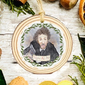 May include: A round wooden Christmas ornament featuring a watercolour illustration of a man holding a gun. The image is framed by a green and red wreath. The text "Yippee Ki-Yay" is on a banner.