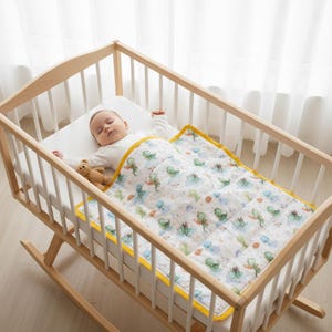 Pode incluir: Um bebé dorme tranquilamente num berço de baloiço de madeira. O bebé está coberto com um edredão branco com um padrão colorido de dinossauros e uma borda amarela. Um pequeno ursinho de pelúcia descansa ao lado do bebé.